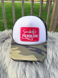 A white and black trucker hat with a camouflage brim, featuring a red square patch on the front with white script and block lettering. 