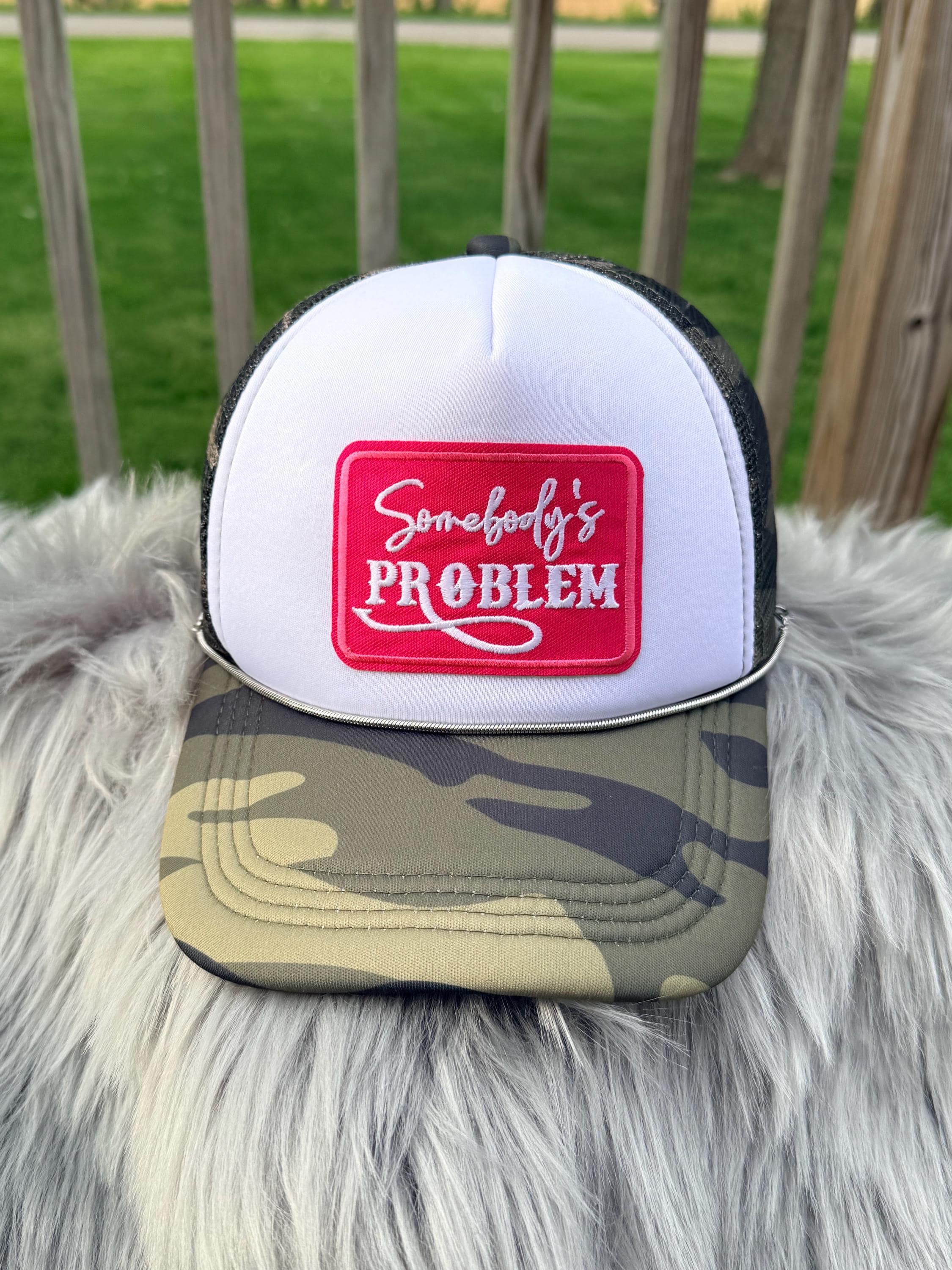 A white and black trucker hat with a camouflage brim, featuring a red square patch on the front with white script and block lettering. 