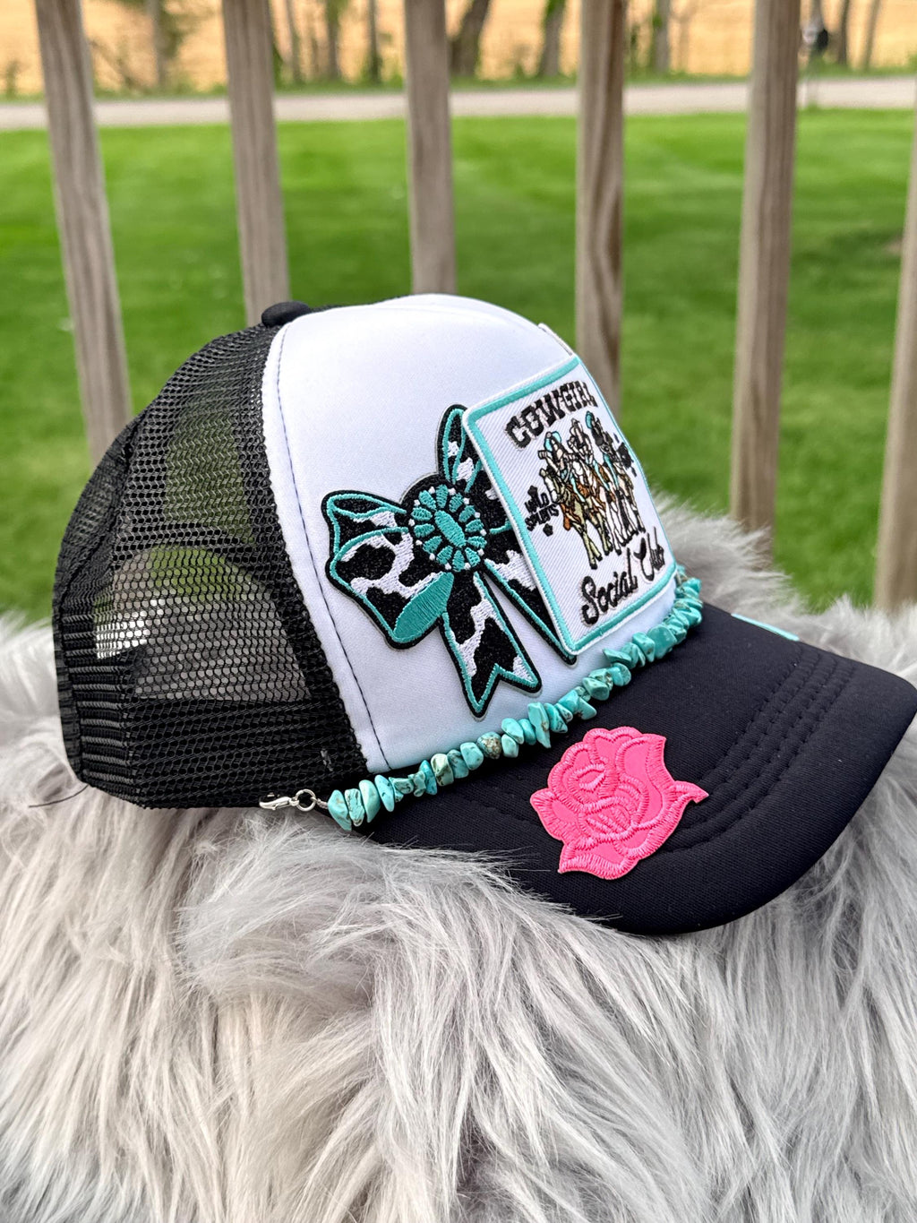 A white and black trucker hat with mesh back panels, decorated with a turquoise and black leopard print bow patch, a 'Country Social Club' embroidered patch, a turquoise beaded trim, and a pink rose patch. 