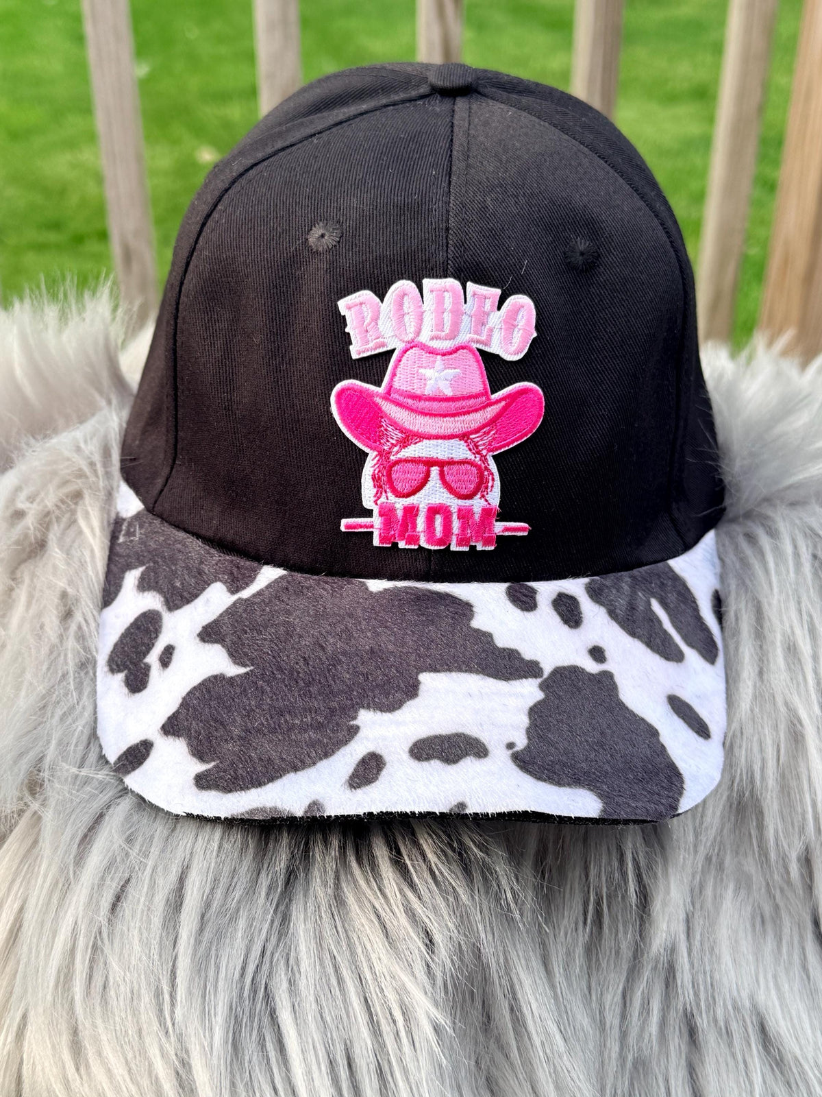 A black baseball cap with a pink embroidered 'RODEO MOM' graphic on the front, featuring a cow-print brim, displayed on a gray faux fur surface with a blurred green outdoor background.