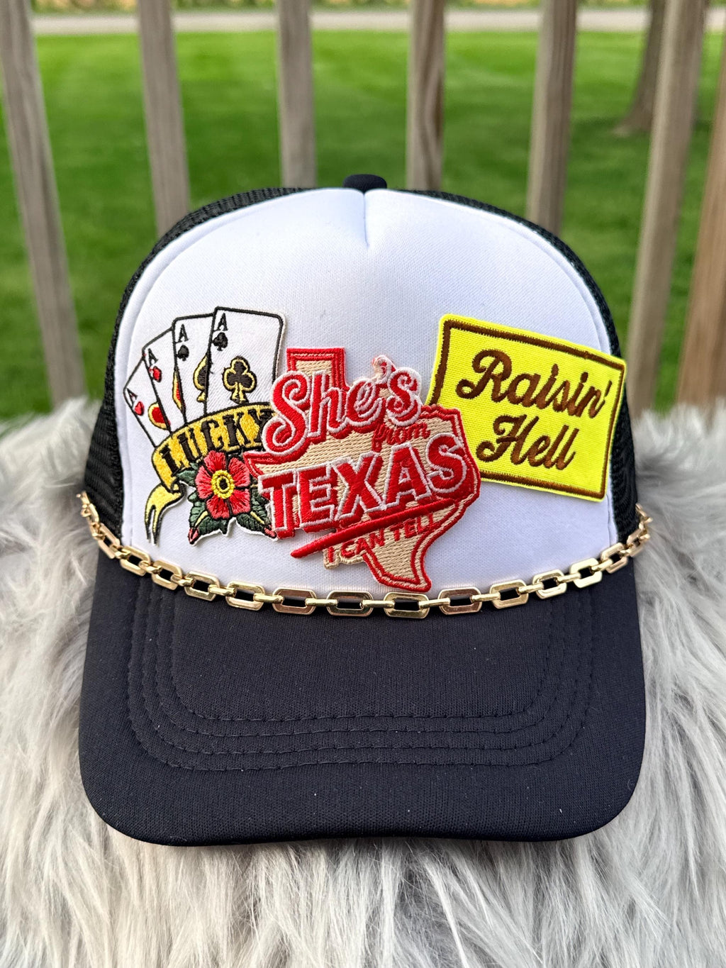 A white and black trucker hat with a gold chain across the brim, featuring embroidered patches including playing cards, a red Texas outline reading ' from Texas and a yellow 'Raisin' Hell' patch, 