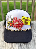A white and black trucker hat with a gold chain across the brim, featuring embroidered patches including playing cards, a red Texas outline reading ' from Texas and a yellow 'Raisin' Hell' patch, 