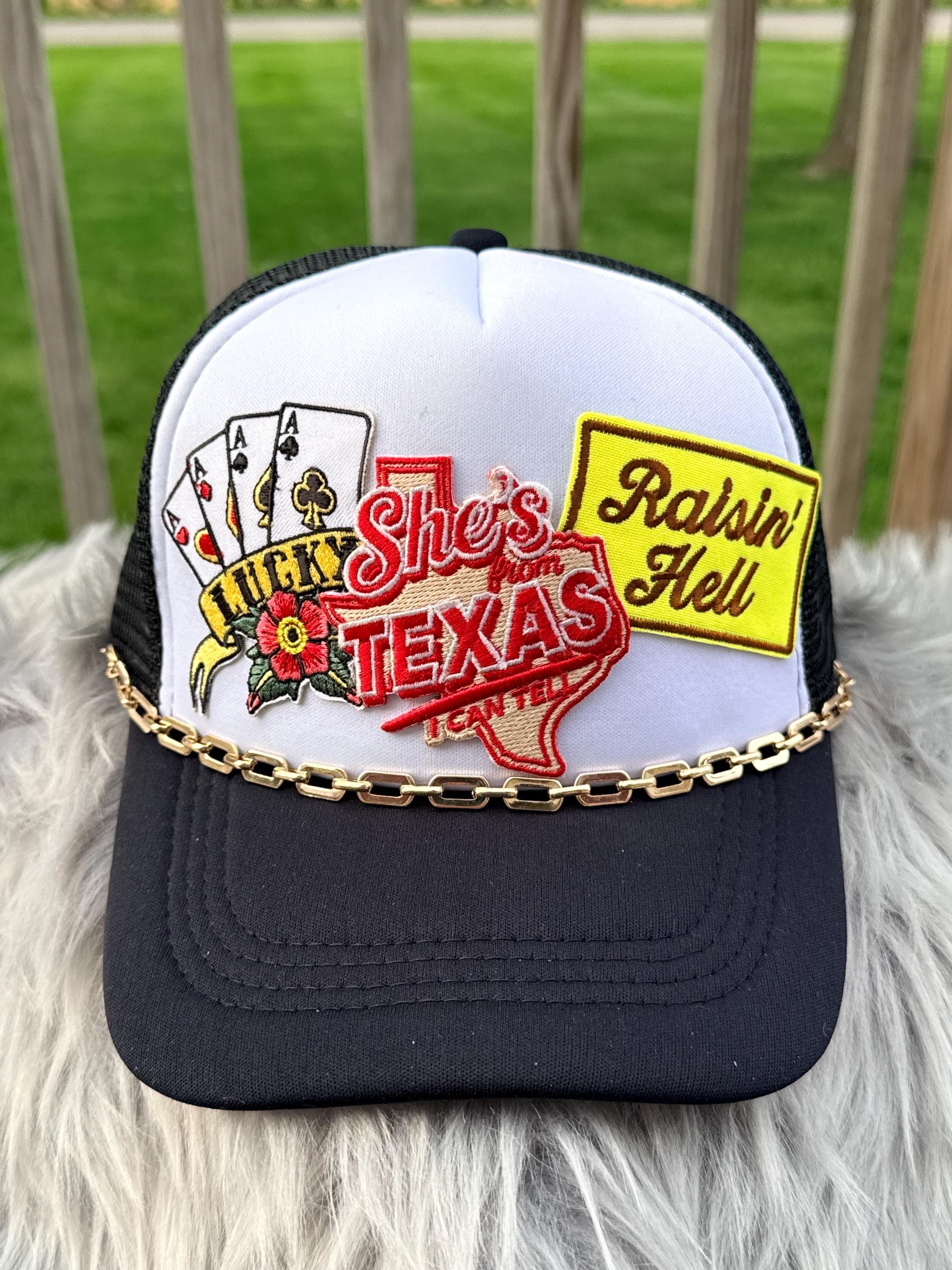 A white and black trucker hat with a gold chain across the brim, featuring embroidered patches including playing cards, a red Texas outline reading ' from Texas and a yellow 'Raisin' Hell' patch, 