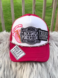A red and white trucker hat with multiple embroidered patches, displayed on a white faux fur surface with a wooden fence and green grass in the background.