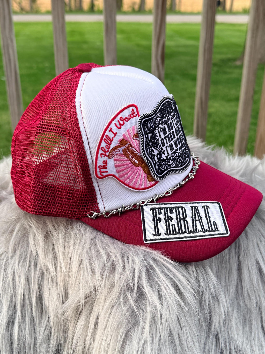 A red and white trucker cap with multiple embroidered patches, including 'The Hell I Won't, a black-and-white crest patch, and a 'FERAL' patch on the brim, resting on a white faux-fur surface with a blurred outdoor background.