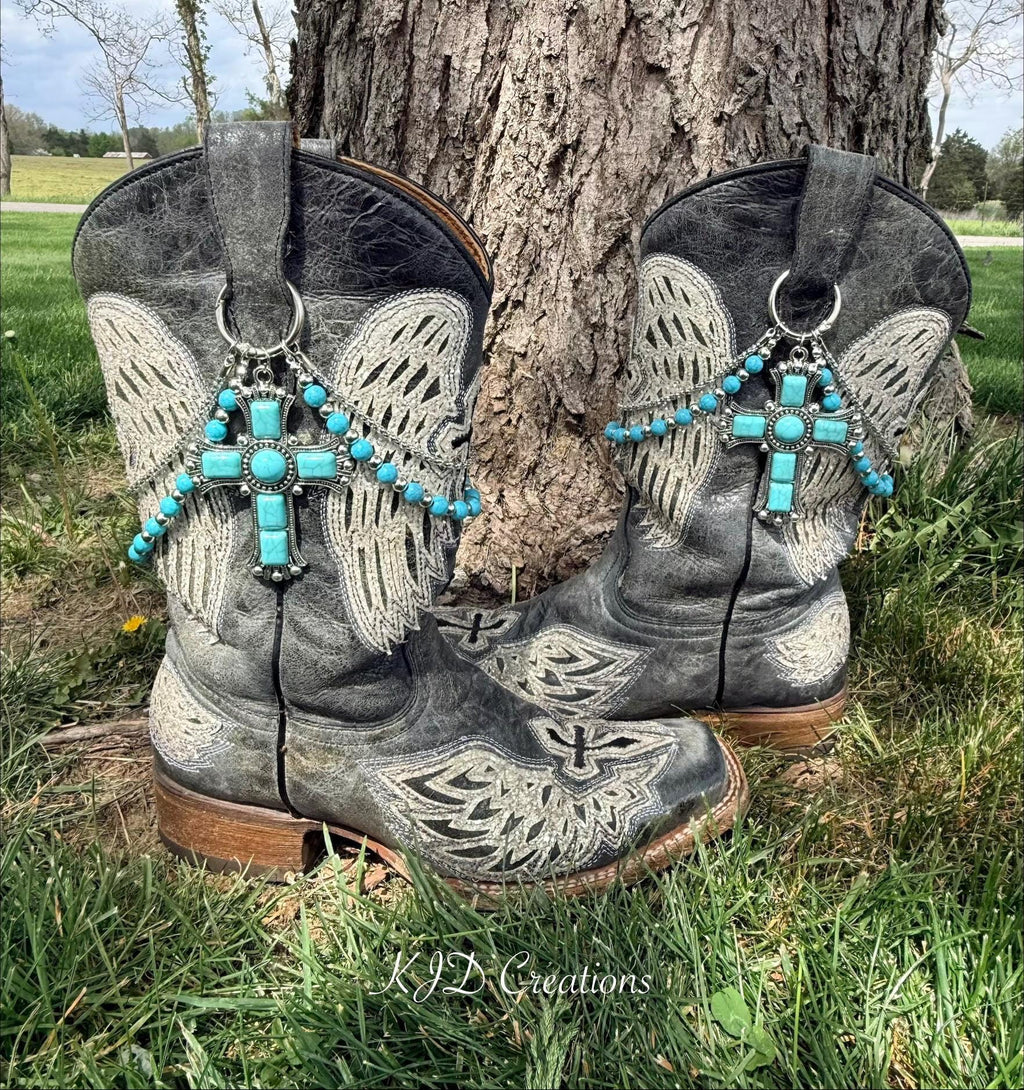 Front view of pair of black distressed angel wing and cross-embroidered cowboy boots on grass against tree wearing two-strand turquoise bead and silver chain lei boot bracelets with massive ornate silver cross pendant set with turquoise cabochon stones, Western boot bling set of 2