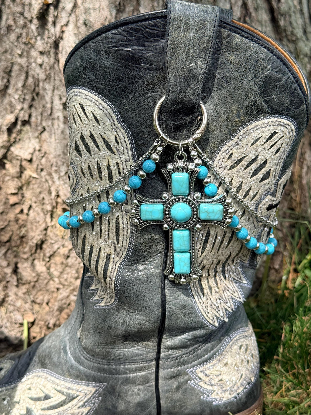 Close-up of black distressed angel wing cowboy boot shaft with two-strand turquoise bead and silver chain lei boot bracelet featuring massive ornate silver cross pendant set with large center oval and rectangular turquoise cabochon stones