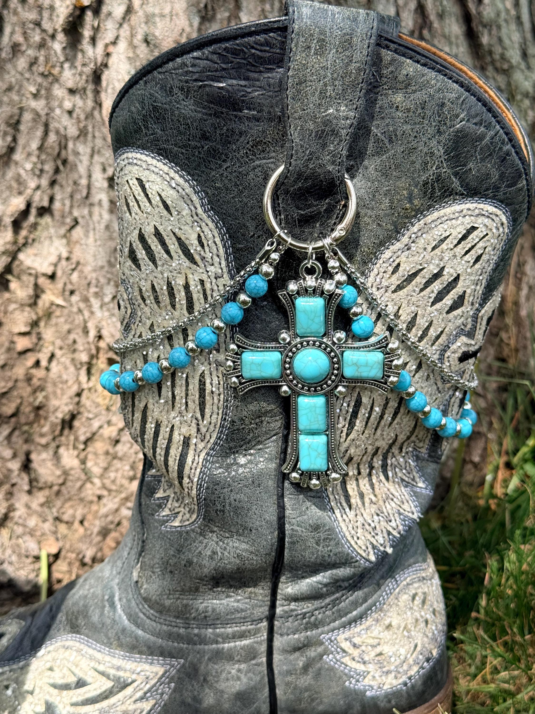 Close-up of black distressed angel wing cowboy boot shaft with two-strand turquoise bead and silver chain lei boot bracelet featuring massive ornate silver cross pendant set with large center oval and rectangular turquoise cabochon stones
