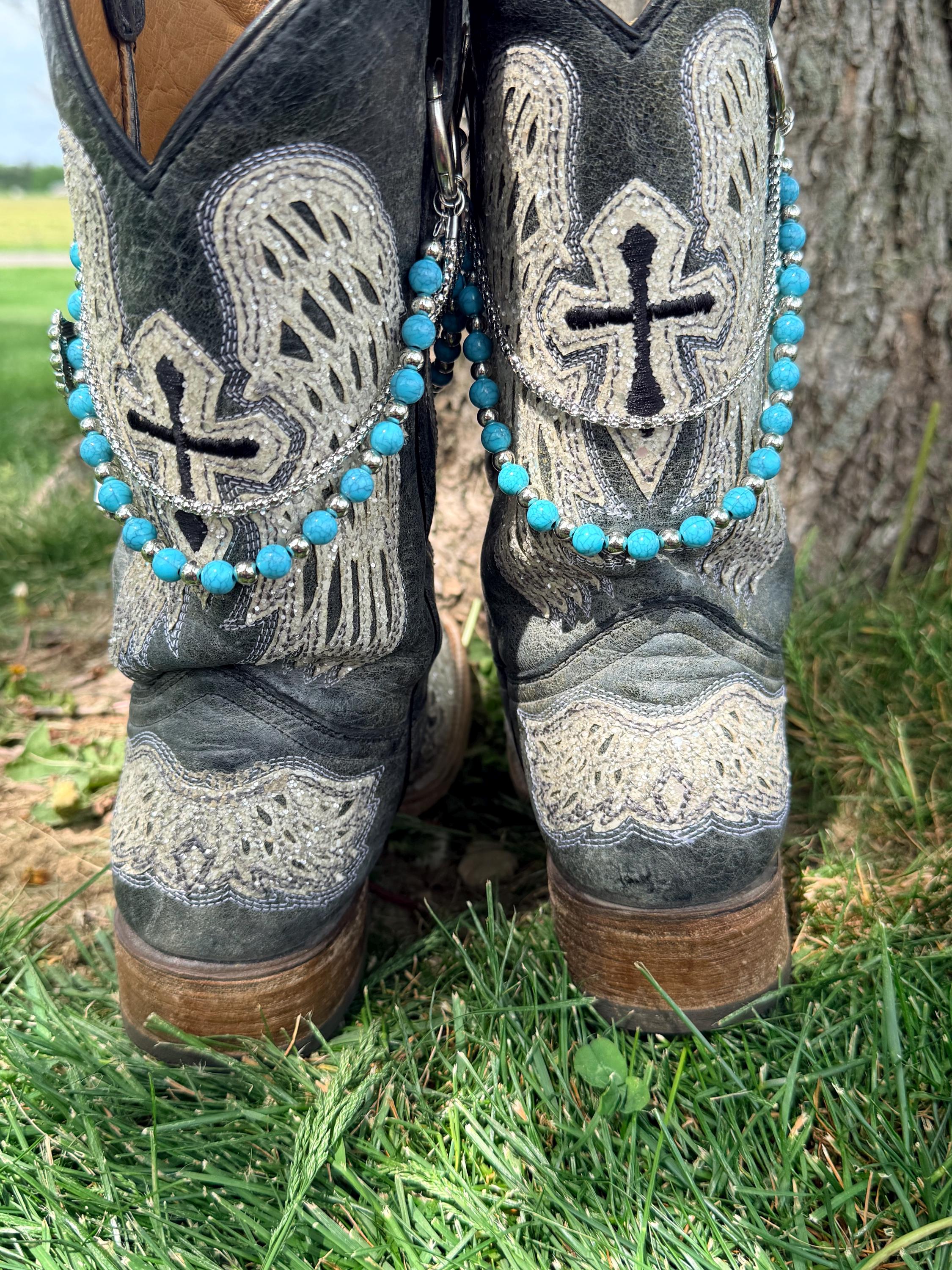 Front view of pair of black cross and angel wing embroidered cowboy boots on grass wearing two-strand turquoise bead and silver chain lei boot bracelets with large silver ring clips through boot pull loop rings, Western boot bling set of 2