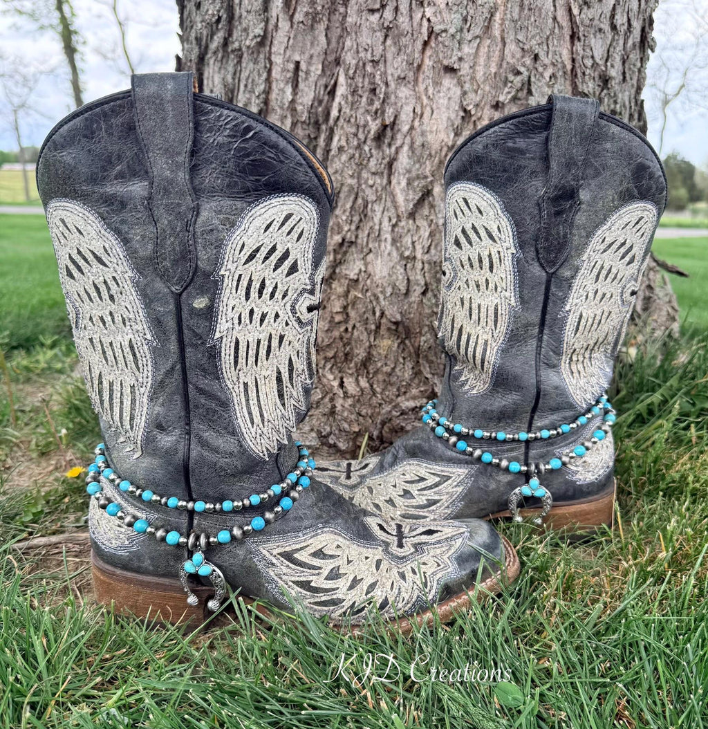 Front view of pair of black distressed angel wing embroidered cowboy boots against tree on grass wearing two-strand turquoise and oxidized silver bead boot bracelets with silver filigree squash blossom naja pendant, Western boho boot jewelry set of 2