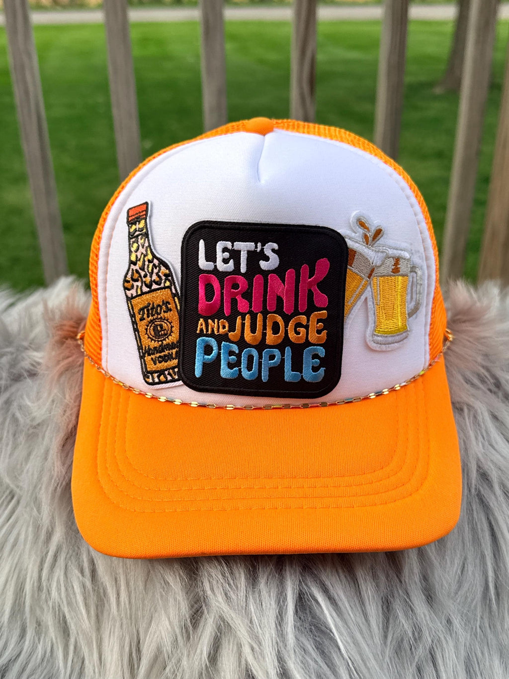 An orange and white trucker hat with a custom embroidered patch that reads 'LET'S DRINK AND JUDGE PEOPLE' alongside a bottle and beer glasses, 
