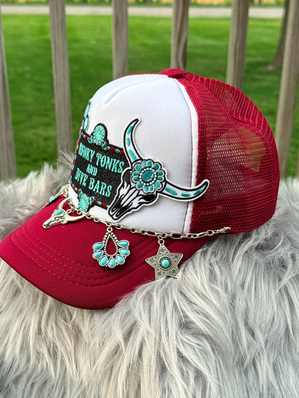 A red and white trucker hat with turquoise and silver western embellishments, including a skull patch, embroidered text, and dangling charms, resting on a white faux fur surface with a wooden fence and green grass in the background.