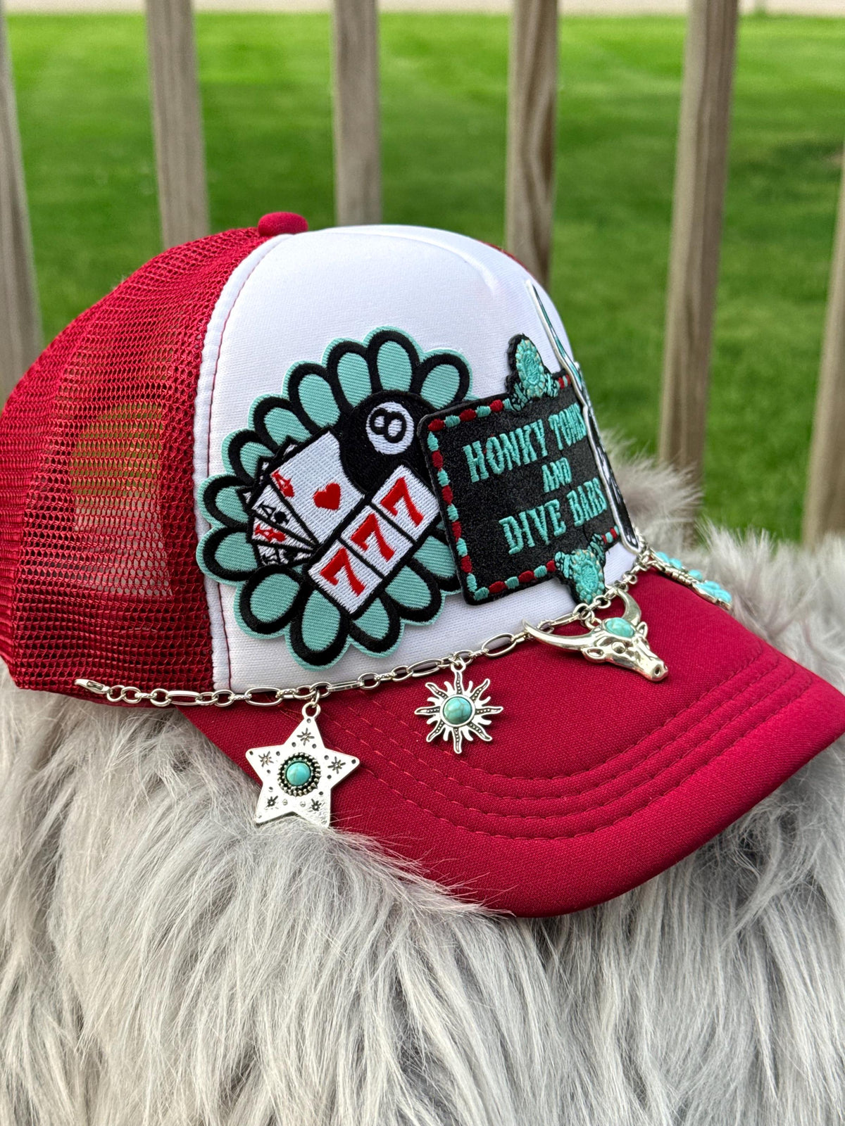 A red and white trucker hat with a white front panel, red mesh back, and multiple decorative patches and charms, including a playing card and 777 patch, a black 'HONKY Tonks' patch, and silver dangling star and sun charms,