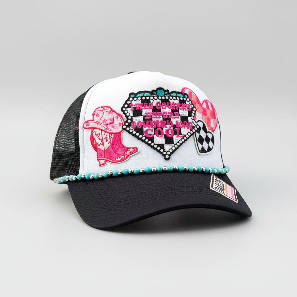 A white and black trucker hat with western-themed patches, including a pink cowboy boot, a checkered heart. The hat has turquoise and silver beads along the brim's edge and a 'DARLIN' brand patch on the side,