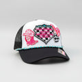 A white and black trucker hat with western-themed patches, including a pink cowboy boot, a checkered heart. The hat has turquoise and silver beads along the brim's edge and a 'DARLIN' brand patch on the side,