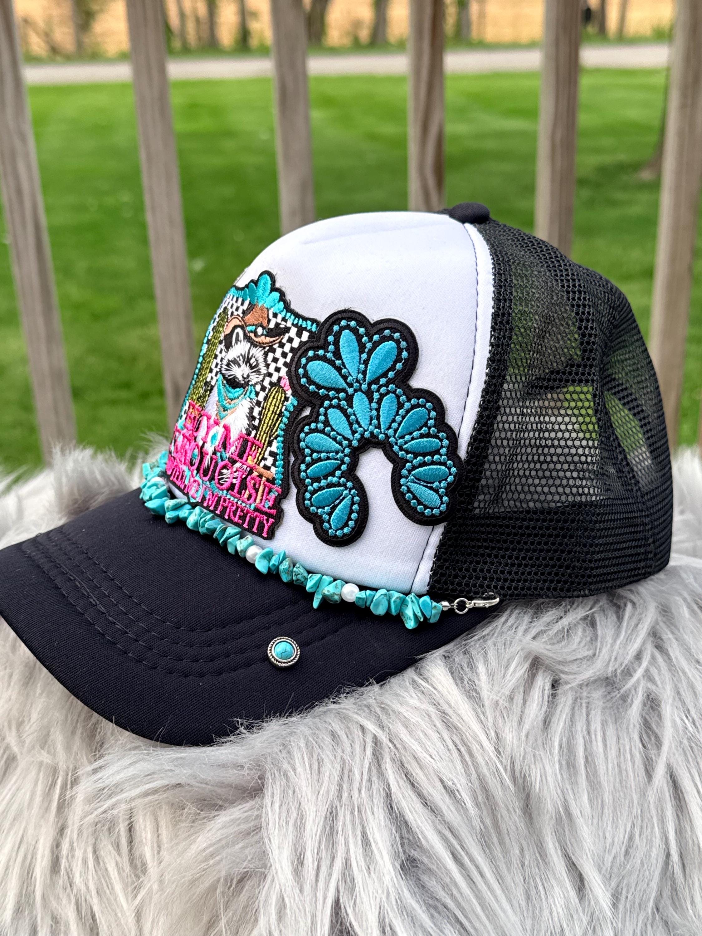 A custom trucker hat with a white front panel, black mesh back, and black brim, decorated with a large turquoise and black floral patch, a colorful embroidered dog design, and turquoise fringe along the edge of the brim. 
