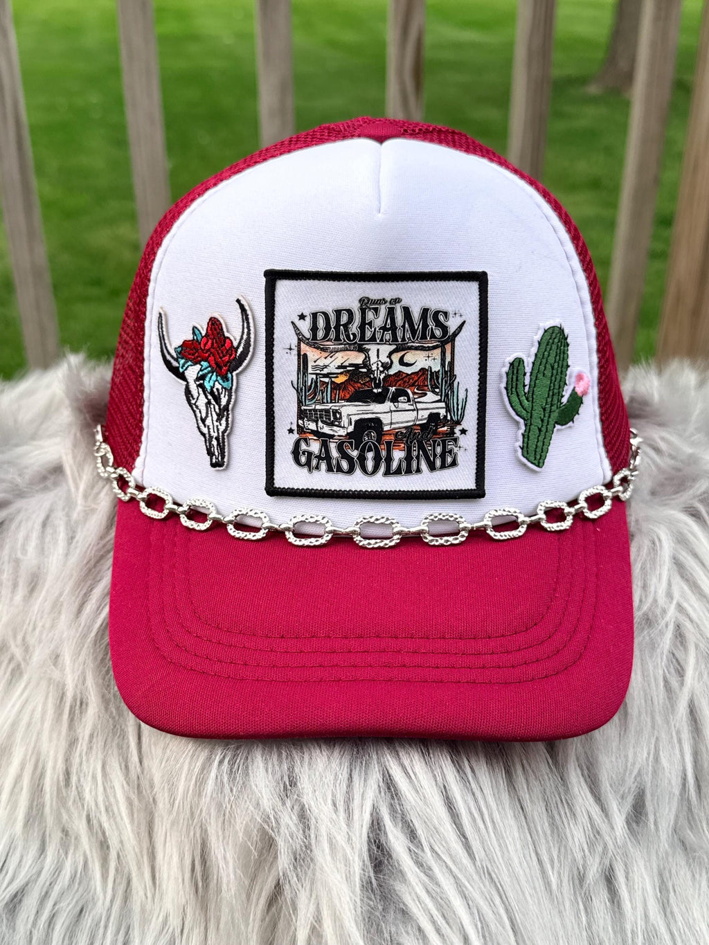 A red and white trucker hat with a silver chain across the front, featuring embroidered patches including a bull skull, a cactus, and a central patch that reads 'Dreams Gasoline'.