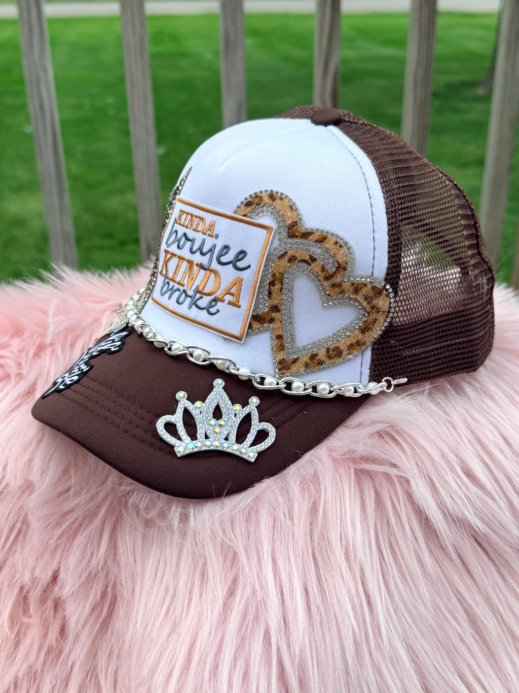 A customized brown and white trucker hat with mesh back, featuring a white front panel with a patch, a leopard print heart outline patch, a silver rhinestone crown patch, and a silver chain across the brim, 