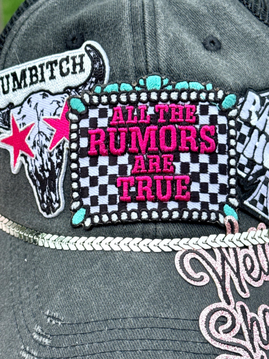 Close-up of a distressed dark gray baseball cap featuring a large embroidered patch that reads 'ALL THE RUMORS ARE TRUE' in pink text over a black-and-white checkered background. Other visible patches include a bull skull with a pink star and partial text on the left, and additional western-style embroidery on the brim.