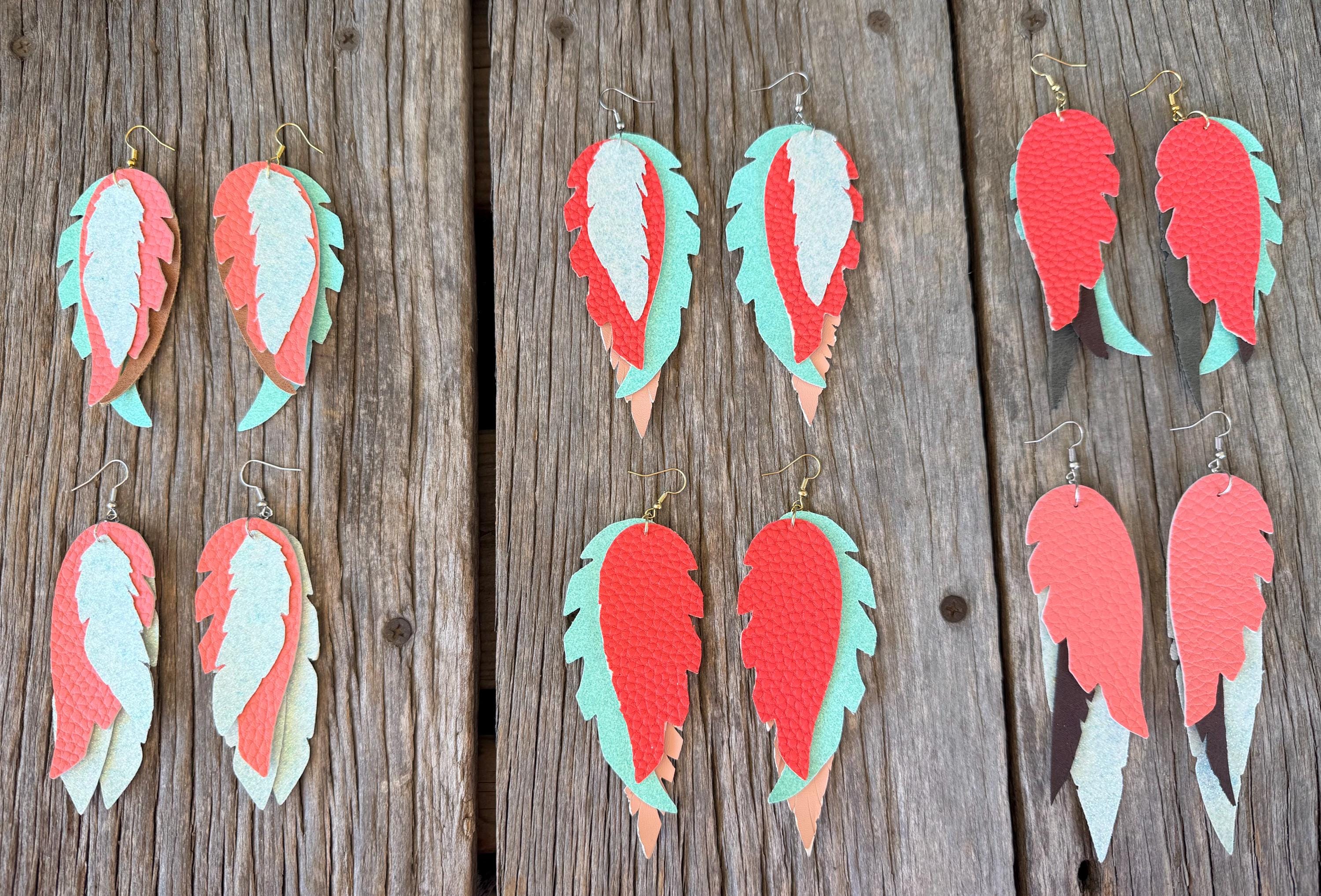 Feather-shaped drop earrings displayed on a rustic wooden plank background. The earrings feature layered feathers in coral pink, mint green, and white, with some showing gold-tone metal hooks.