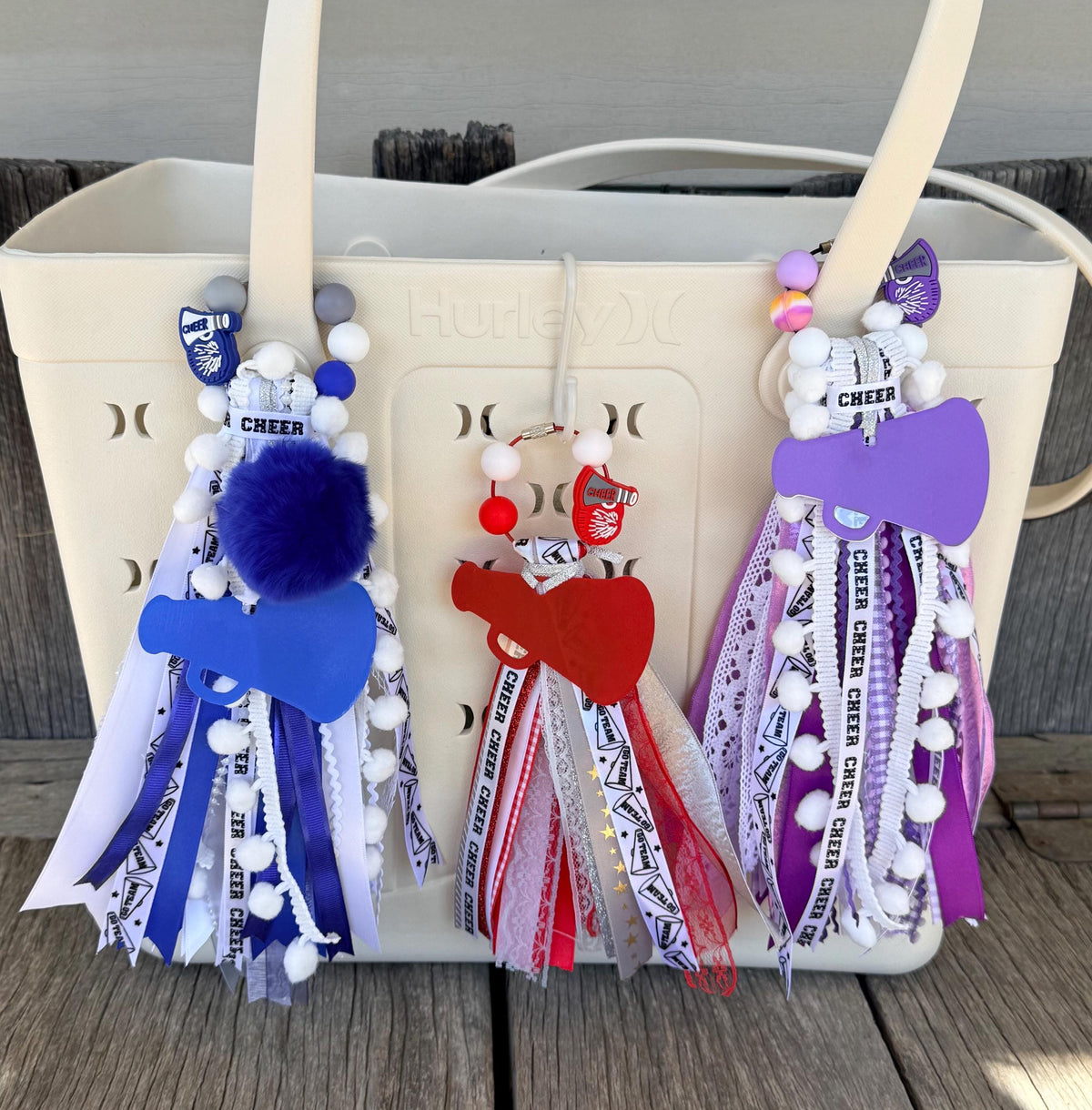 A cream-colored Hurley bag with three decorative cheer-themed tassel charms hanging from the handles, set on a wooden deck.
