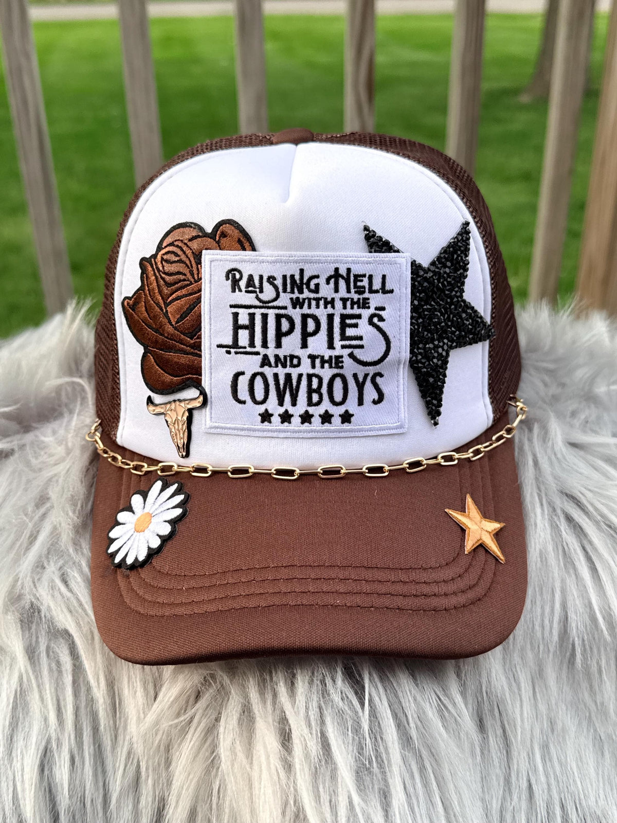 A brown and white trucker hat with mesh back, decorated with a brown ice cream cone patch, a black glitter star, a white daisy patch, a gold chain across the brim, and a gold star patch. The front patch reads 'RAISING HELL WITH THE HIPPIES AND THE COWBOYS'.