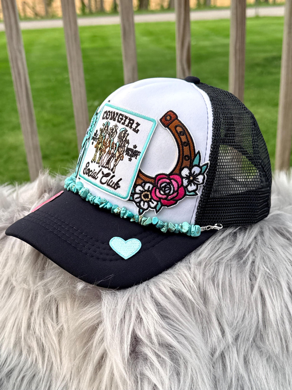 A white and black trucker hat with a 'Cowgirl Social Club' patch, turquoise beaded trim, a horseshoe and rose graphic, and a light blue heart on the brim, resting on a white faux fur surface with a blurred green yard in the background.