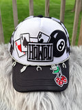 A trucker-style baseball cap with a white front panel and black brim, decorated with patchwork graphics including playing cards, an 8-ball, the word 'HOWOU', and hanging silver charms. 