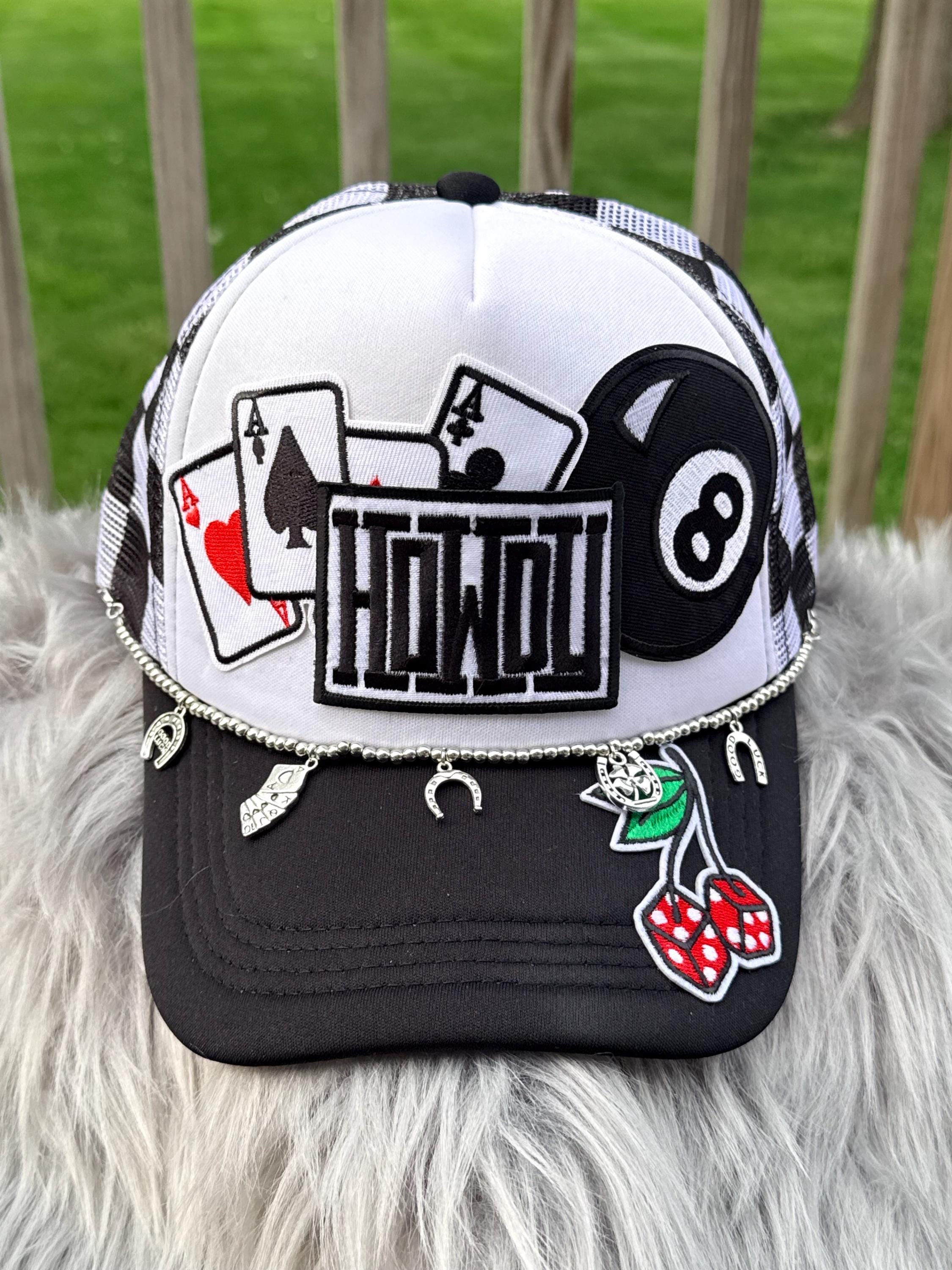 A trucker-style baseball cap with a white front panel and black brim, decorated with patchwork graphics including playing cards, an 8-ball, the word 'HOWOU', and hanging silver charms. 