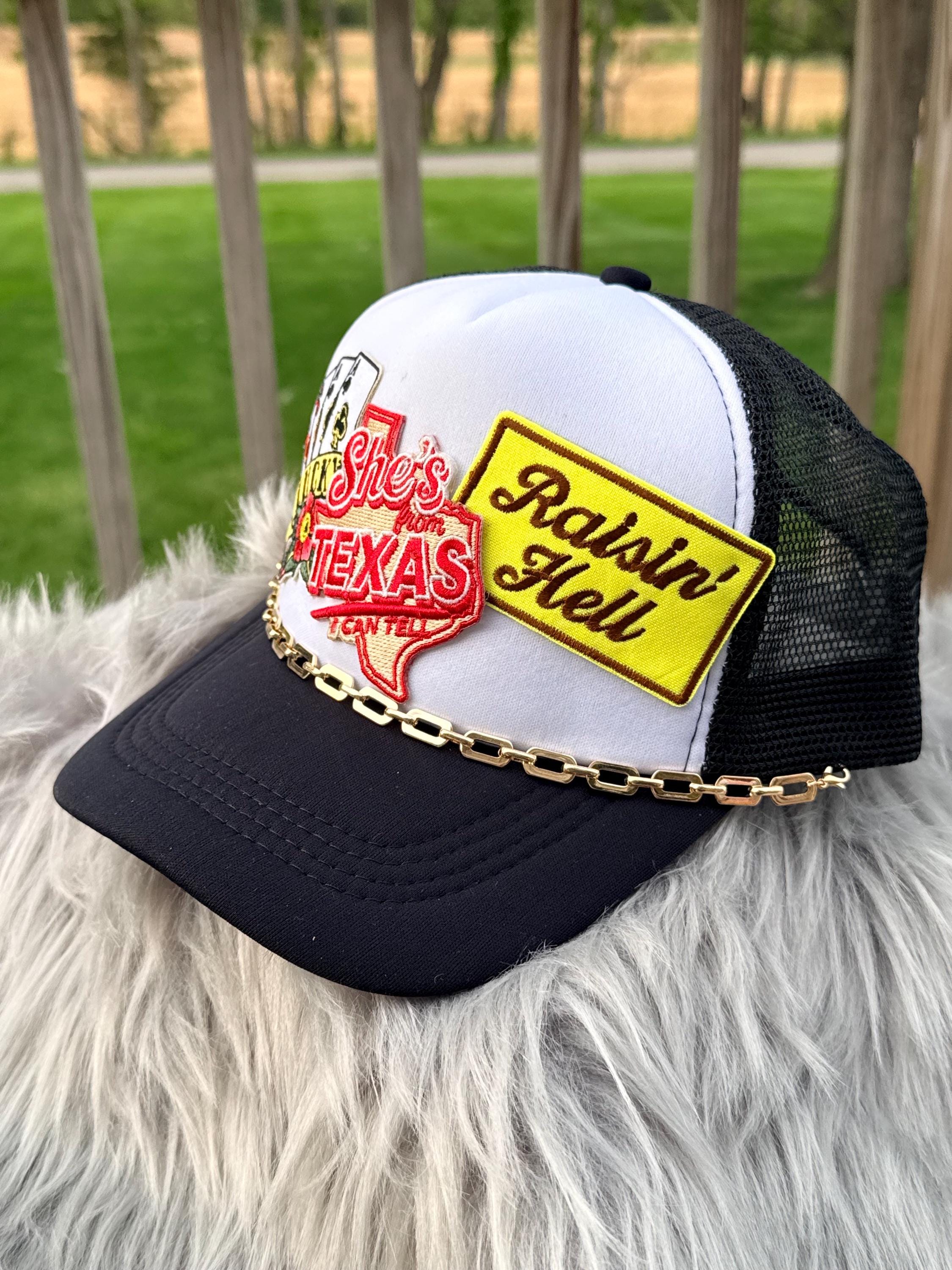 A black and white trucker hat with mesh back panels, featuring a white front panel with a red and white Texas-shaped patch  and a yellow patch reading 'Ratsin' Hell,' plus a gold chain draped across the brim. T