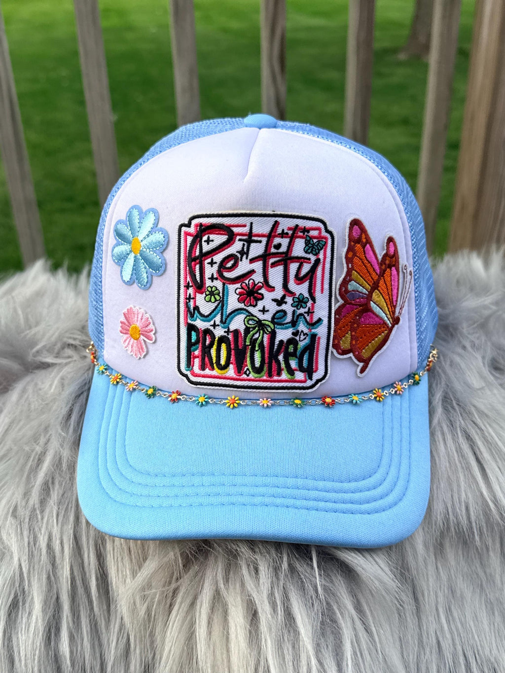 A light blue and white trucker hat with colorful embroidered details, including flowers, a butterfly, and the text 'Pretty When Provoked,'