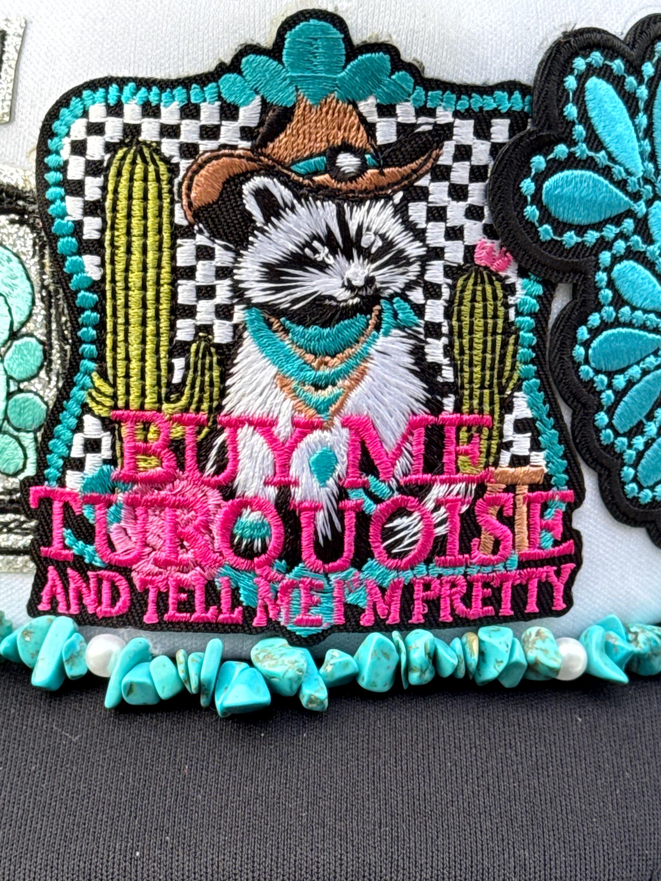 A close-up of a decorative embroidered patch featuring a raccoon in a cowboy hat with cacti and turquoise accents, plus the text 'BUY ME TURQUOISE AND TELL ME I'M PRETTY,' shown on a dark fabric background.