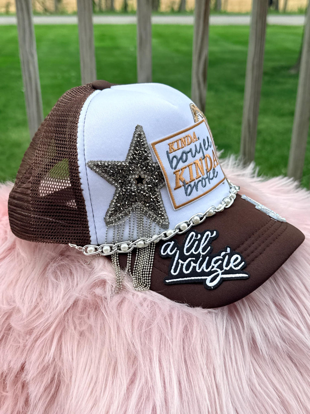 A brown and white trucker-style cap with mesh back panels, embellished with a large rhinestone star, a patch reading 'KINDA boujie KINDA broke,' a silver chain detail, and white script embroidery on the brim that reads 'a lil bougie.' 