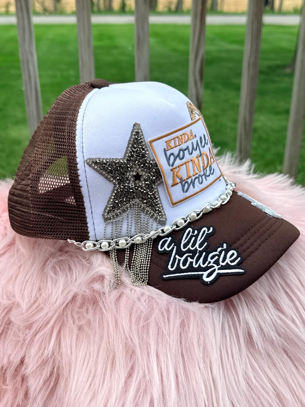 A brown and white trucker-style cap with mesh back panels, embellished with a large rhinestone star, a patch reading 'KINDA boujie KINDA broke,' a silver chain detail, and white script embroidery on the brim that reads 'a lil bougie.' 