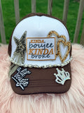 A brown and white trucker hat with 'KINDA boujee KINDA broke' embroidered on the front, decorated with rhinestone star, heart, and crown patches, a rhinestone chain, and a 'a lil Bougie' patch on the brim, 