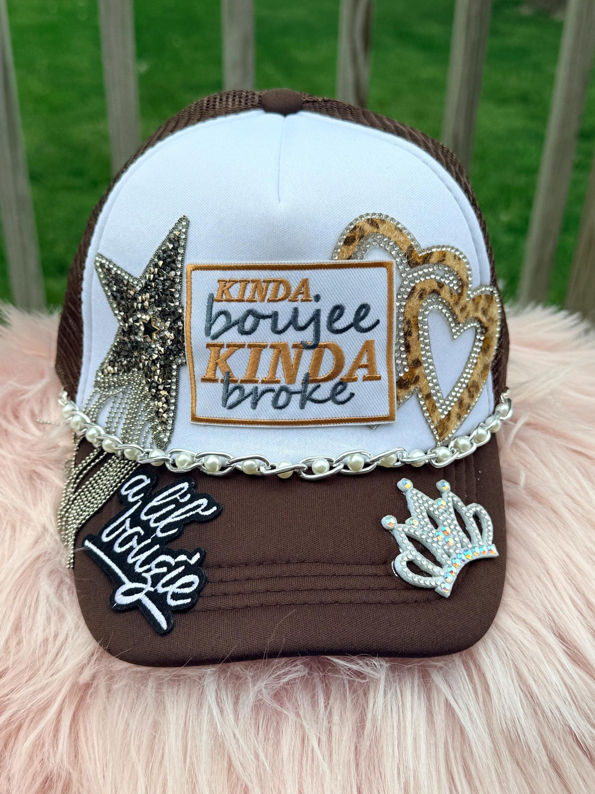 A brown and white trucker hat with 'KINDA boujee KINDA broke' embroidered on the front, decorated with rhinestone star, heart, and crown patches, a rhinestone chain, and a 'a lil Bougie' patch on the brim, 