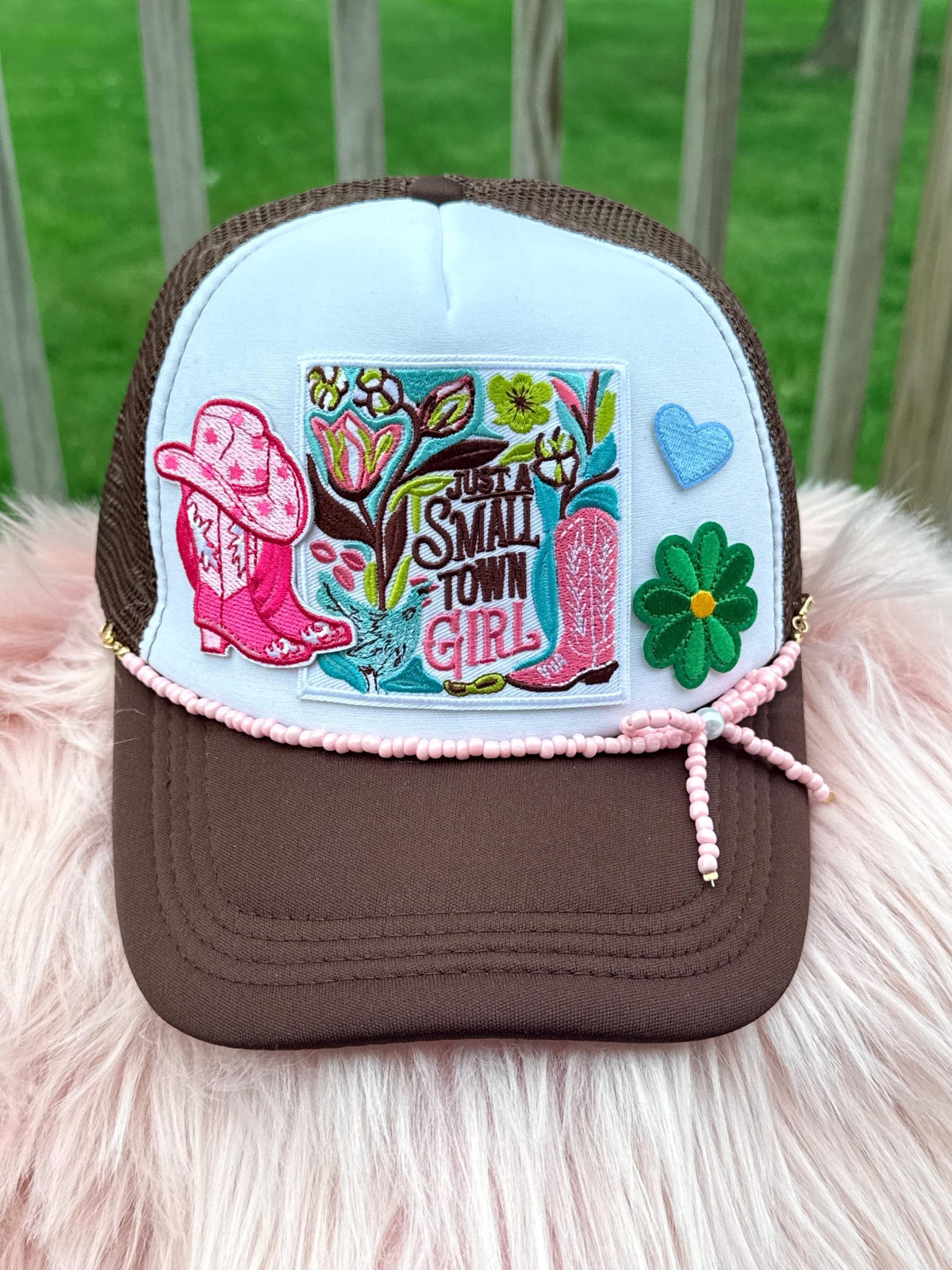 A brown and white trucker hat with a colorful embroidered patch reading ' Small Town Girl,' pink cowboy boots, a green flower, and a blue heart,