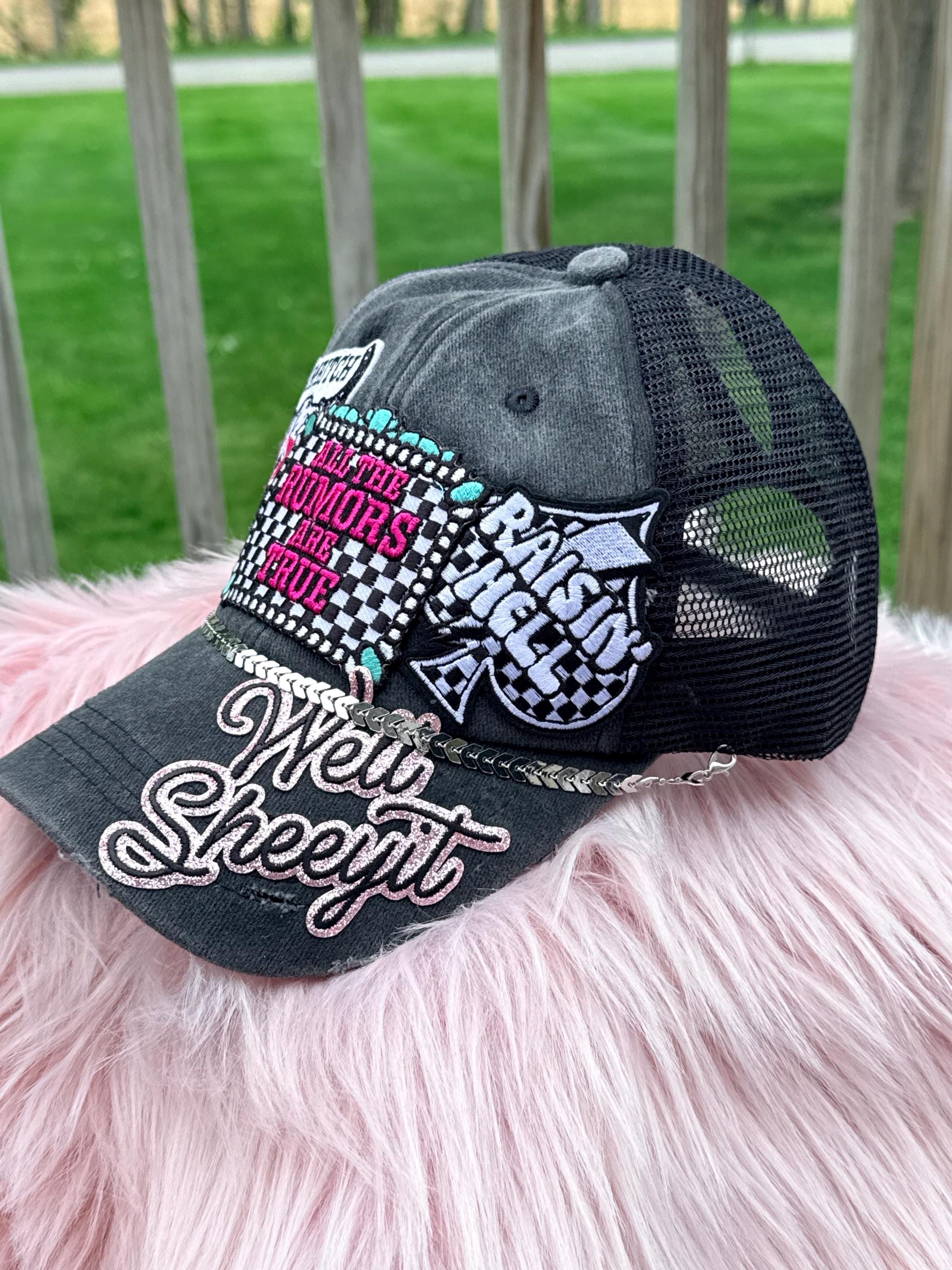 A distressed black trucker cap with mesh back panels, featuring multiple embroidered patches and rhinestone details on the front and brim. 