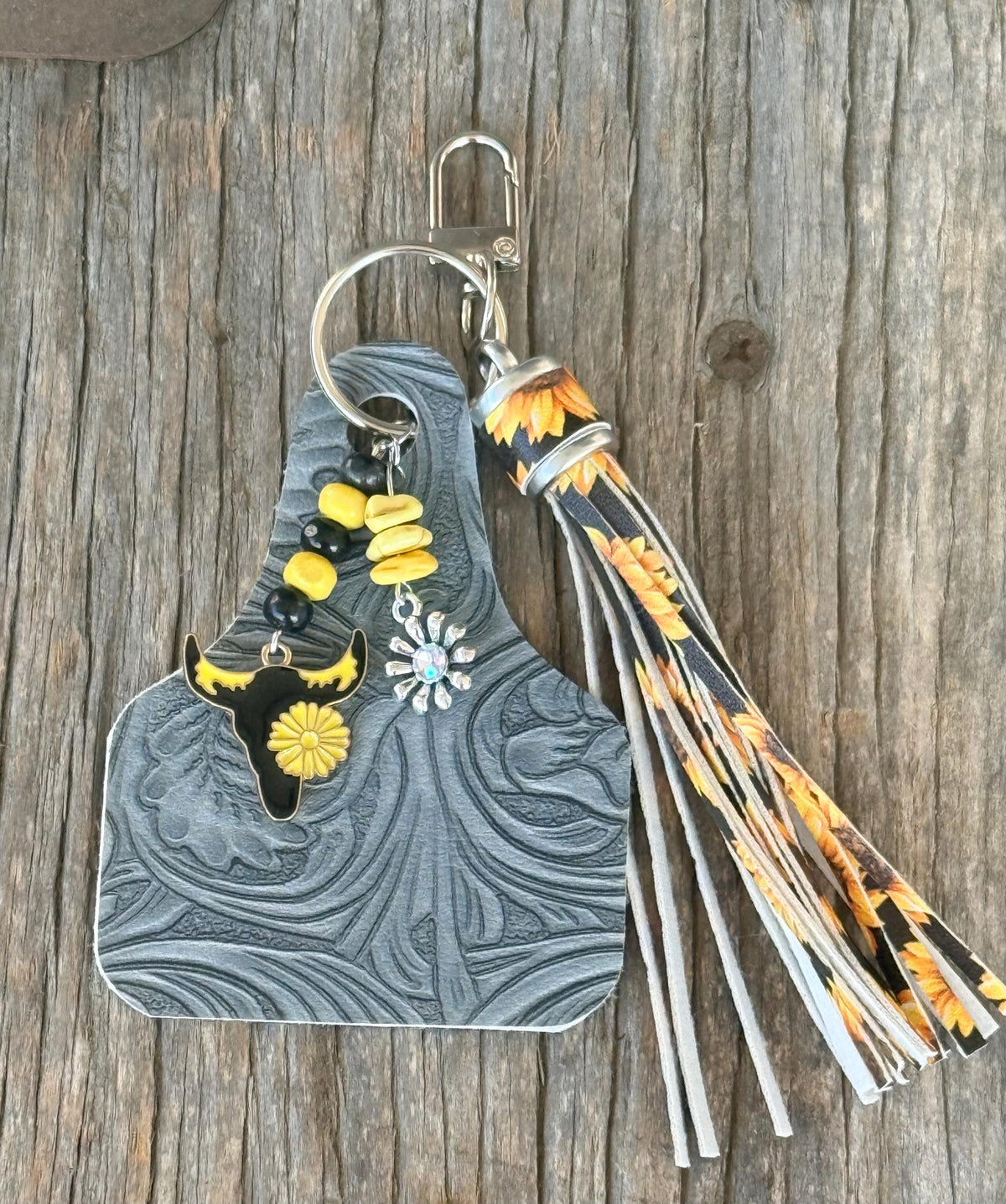 Flat lay of gray-blue tooled leather cow tag keychain bag charm with sunflower print fabric fringe tassel, yellow turquoise chip beads, black enamel longhorn skull with yellow sunflower charm, and silver daisy concho on silver lobster clasp, on weathered wood