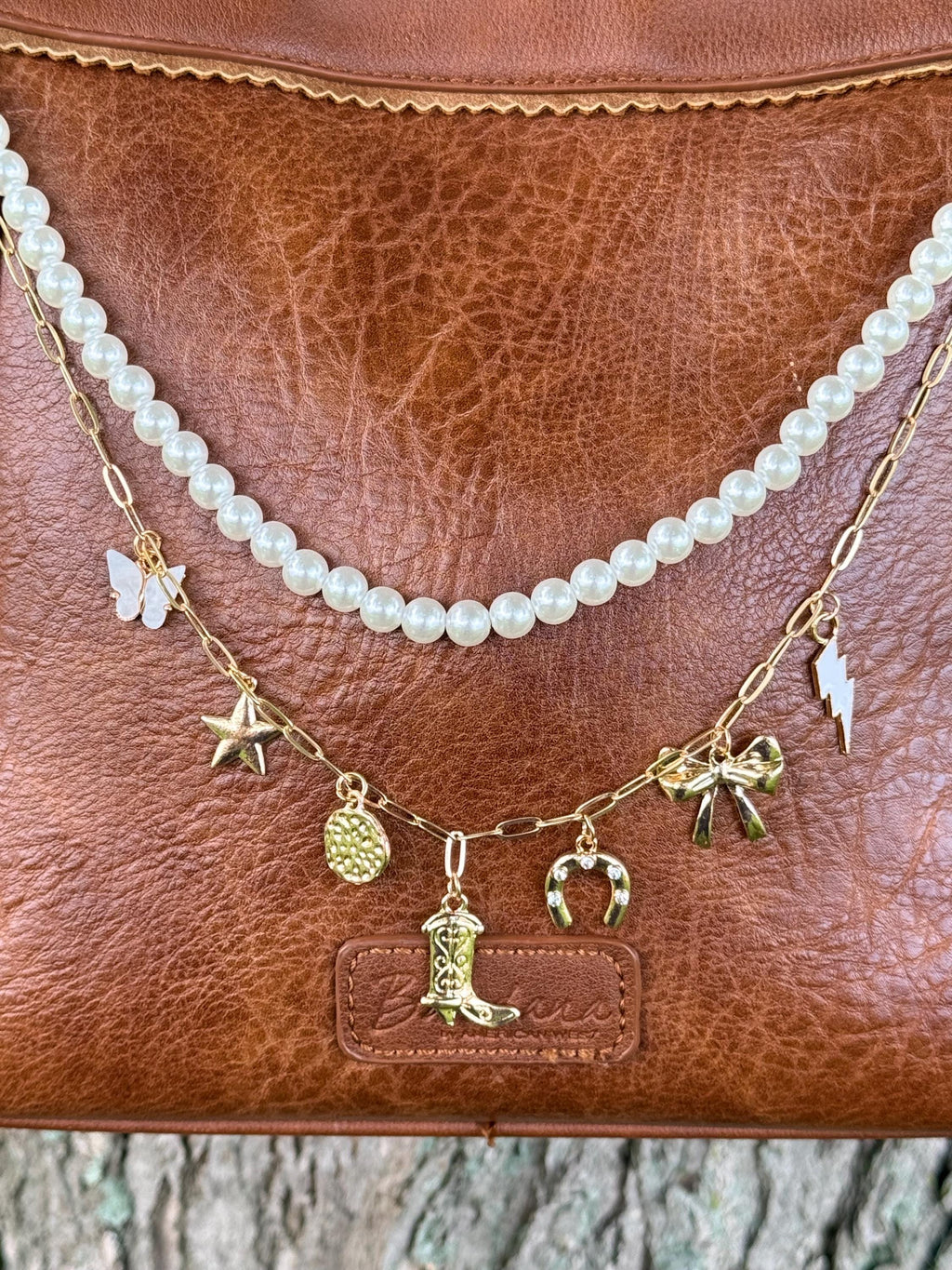 Close-up of brown distressed leather handbag with two-strand Western boho purse chain showing faux pearl strand and gold paperclip link chain with seven gold Western charms including cowboy boot, horseshoe, star, bow, lightning bolt, coin medallion, and white butterfly