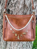 Front view of brown distressed leather crossbody handbag on tree with two-strand Western boho purse chain featuring faux pearl strand and gold paperclip link chain with seven gold charms: white butterfly, star, coin medallion, cowboy boot, horseshoe, bow, and lightning bolt