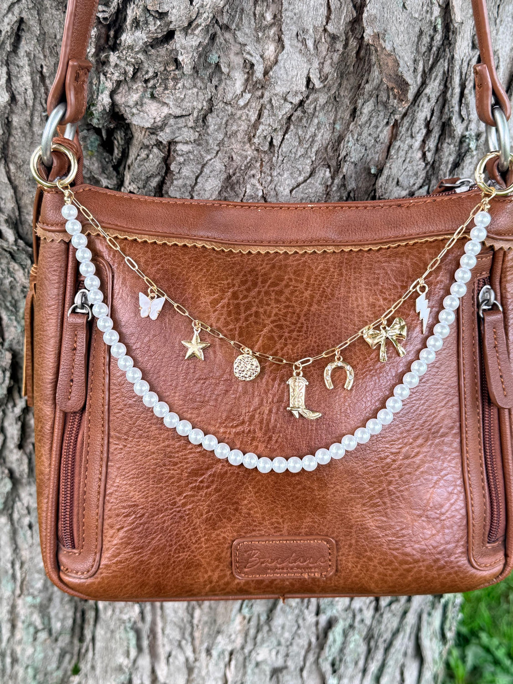 Front view of brown distressed Bandana leather crossbody bag against tree bark with two-strand Western boho purse chain featuring gold paperclip link chain with seven Western charms including white enamel butterfly, gold star, gold coin medallion, gold cowboy boot, gold horseshoe, gold bow, and gold lightning bolt, with faux pearl bead strand draping lower, 11-inch Western handbag necklace