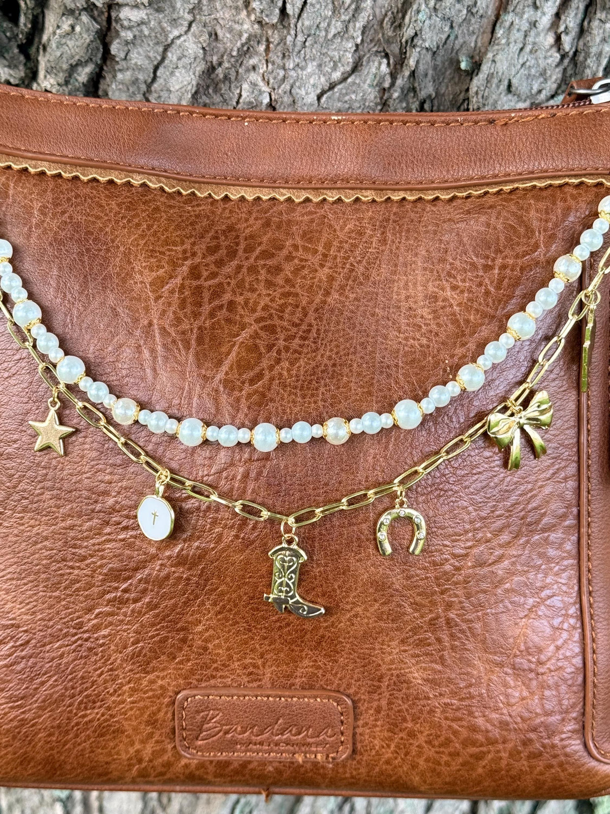 Close-up of brown distressed Bandana leather crossbody bag with gold paperclip link chain featuring five Western charms: gold star, white enamel oval cross medallion, ornate gold cowboy boot, gold horseshoe, and gold bow, with faux pearl bead strand with mixed-size pearls above, Western boho 12.5-inch purse chain detail