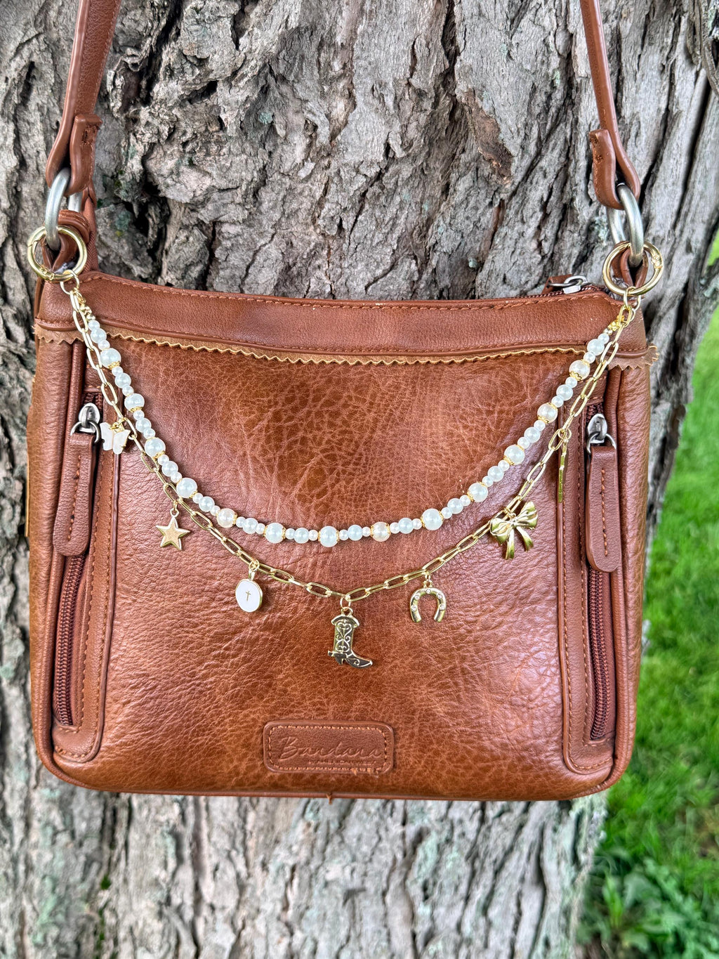 Front view of brown distressed Bandana leather crossbody bag against tree with two-strand Western boho purse chain featuring gold paperclip link chain with five Western charms including gold star, white enamel oval cross medallion, ornate gold cowboy boot, gold horseshoe, and gold bow, plus faux pearl bead strand with mixed-size pearls, 12.5-inch handbag jewelry