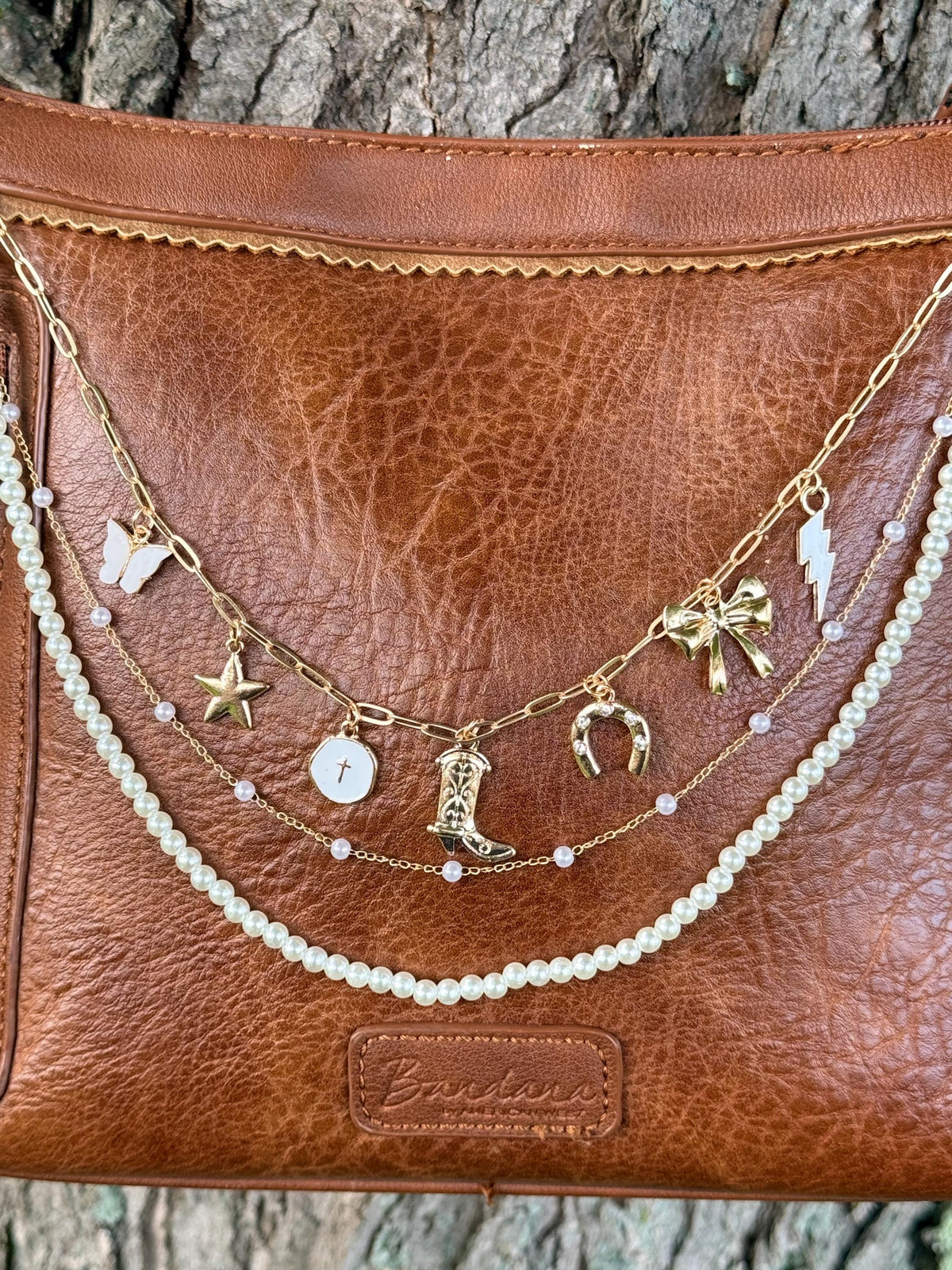 Close-up of brown leather Bandana crossbody bag with gold paperclip link chain featuring seven Western charms: white butterfly, gold star, white cross coin disc, ornate cowboy boot, gold horseshoe, gold bow, and gold lightning bolt, with delicate gold chain with pink bead accents and faux pearl bead strand, 13-inch Western boho purse chain
