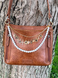 Front view of brown distressed Bandana leather crossbody bag against tree with three-strand Western boho purse chain featuring gold paperclip link chain with eleven cheetah print and gold charms, gold crystal rhinestone tennis chain, and faux pearl bead strand, 10-inch Western handbag jewelry