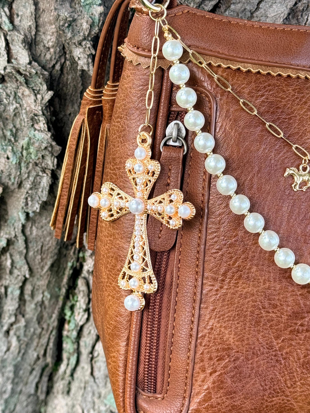 Close-up of large ornate gold filigree cross pendant set with pearl cabochons hanging from brown leather Bandana crossbody bag zipper hardware alongside gold leather fringe tassel, with faux pearl bead strand and gold paperclip link chain visible, Western boho purse chain detail
