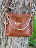Front view of brown leather Bandana crossbody bag against tree with two-strand Western boho purse chain featuring gold paperclip link chain with seven Western charms, faux pearl bead strand, large ornate gold filigree cross pendant set with pearl cabochons, and gold leather fringe tassel