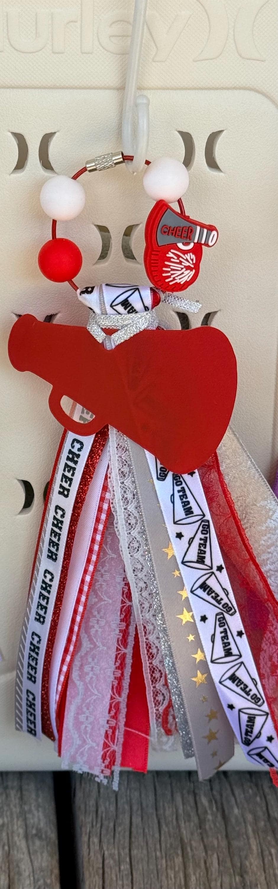 Alternate close-up of red personalized cheer silicone beaded bag tassel clipped to white Hurley tote bag handle showing large red acrylic megaphone-heart pendant with personalized name, silver glitter ribbon bow, and cascading red and white Cheer print grosgrain white lace red lace silver glitter and Go Team print ribbon tassel, KJD Creations