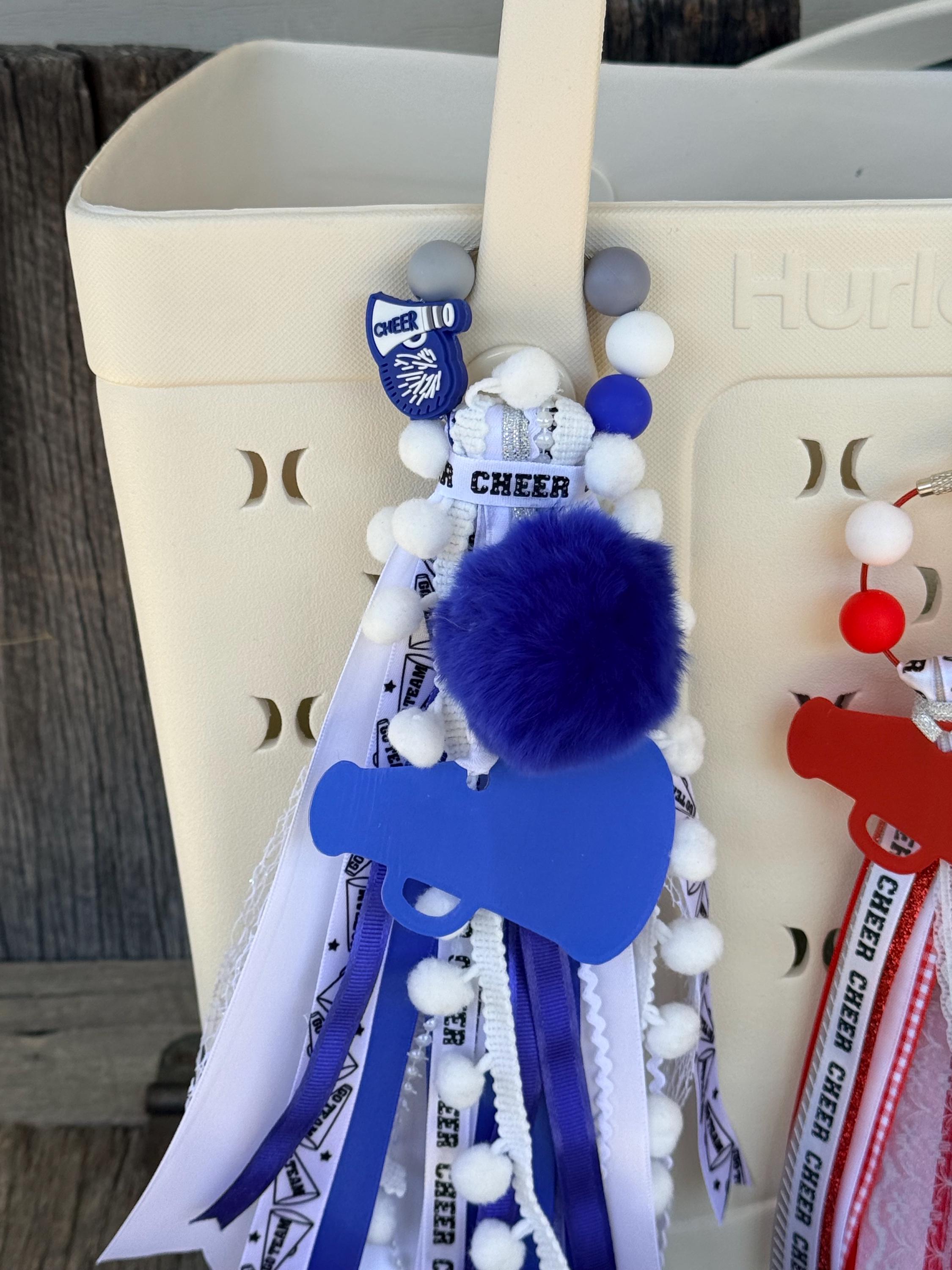 Close-up of blue personalized cheer silicone beaded bag tassel clipped to white Hurley tote bag handle showing large blue acrylic megaphone pendant with blue faux fur pom-pom, silver glitter CHEER ribbon bow, and cascading blue and white Cheer print grosgrain and white pom-pom trim ribbon tassel, with red variant partially visible at right, KJD Creations cheerleading bag charm